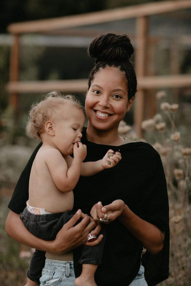 Black woman with braids up in a bun wearing a black Cocoon breastfeeding poncho and holding a shirtless baby with curls