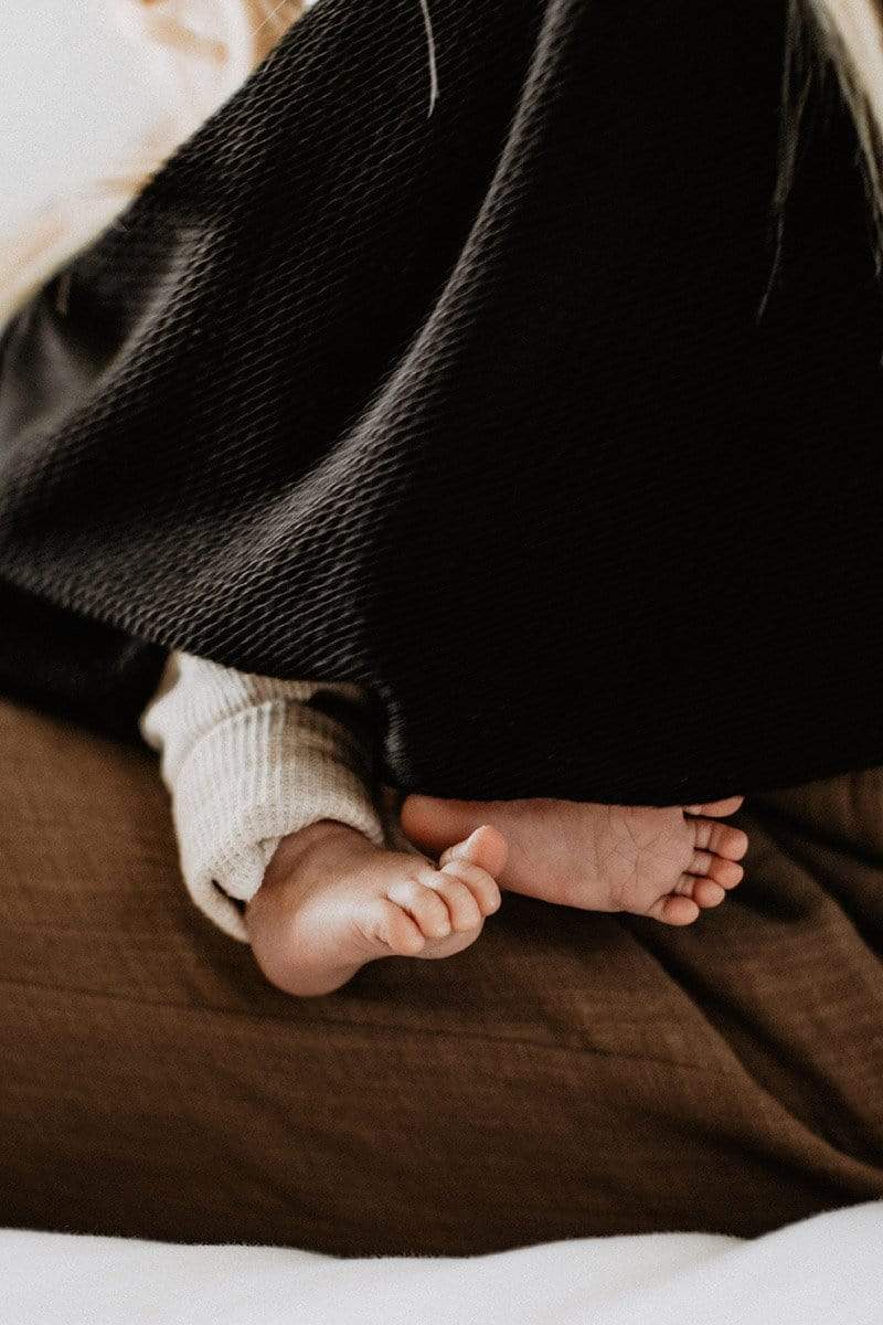 Side view closeup of a mother in brown linen pants breastfeeding under a black knit nursing cover with baby toes peeking out from underneath