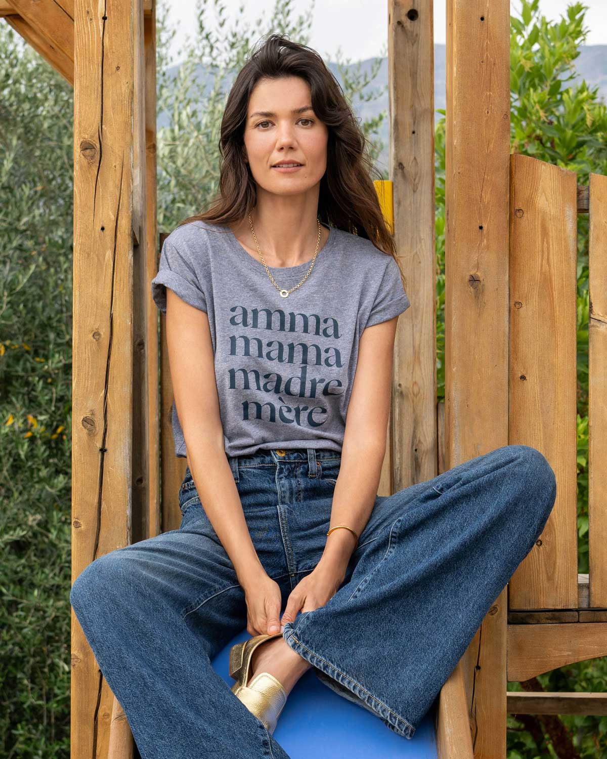 Young mom with long brown hair sitting on slide wearing a heathered grey tee shirt that reads Amma, Mama, Madre, Mere