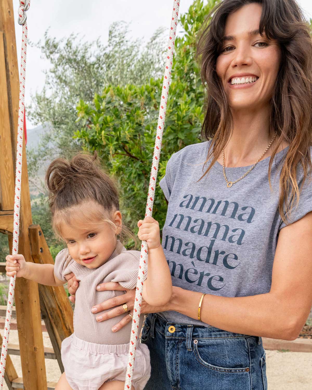 Young mom with long brown hair pushing her toddler on a swingset wearing a heathered grey tee shirt that reads Amma, Mama, Madre, Mere