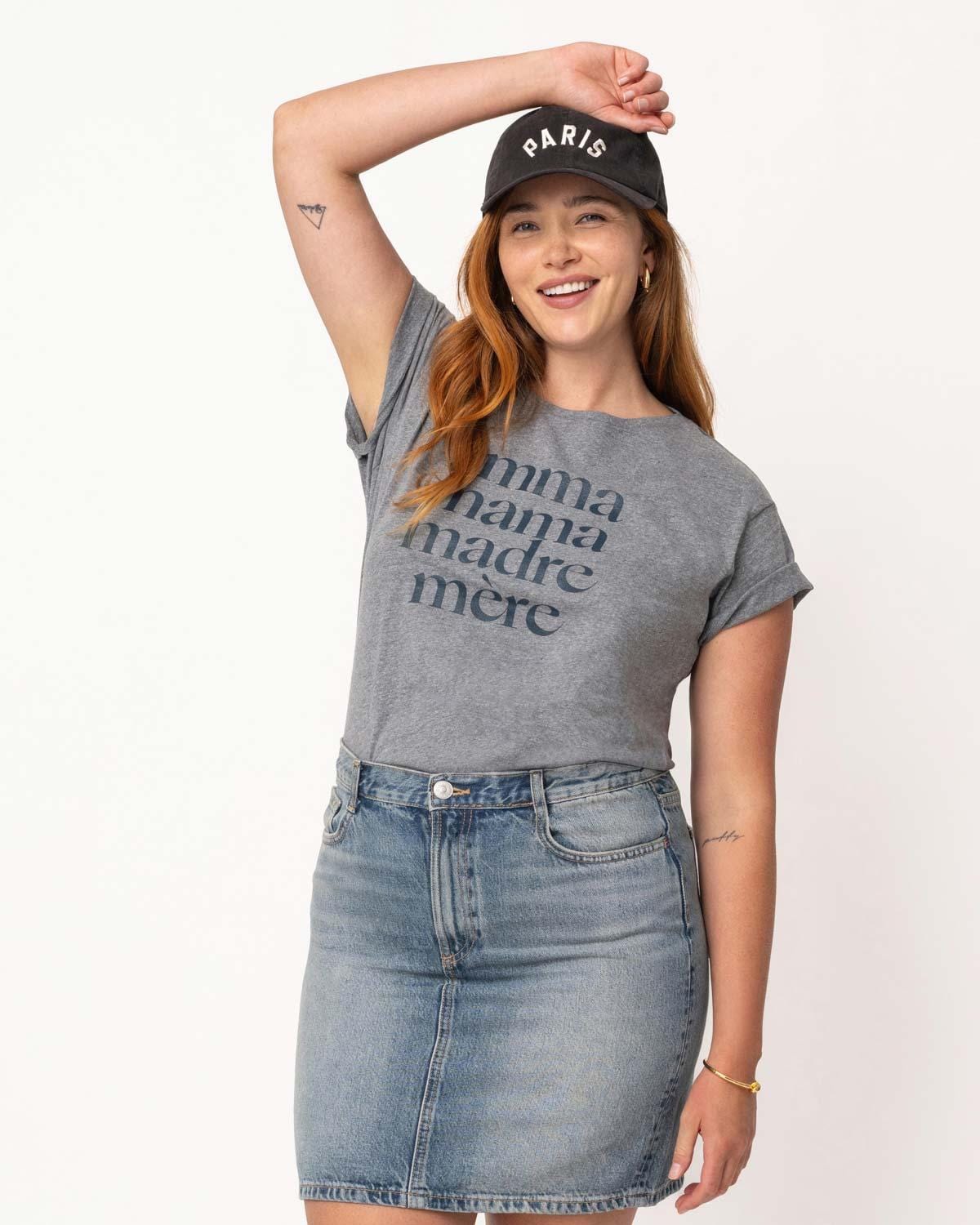 Young mom with long red hair wearing a ball cap, denim skirt and a heathered grey tee shirt that reads Amma, Mama, Madre, Mere in size XL on a white background