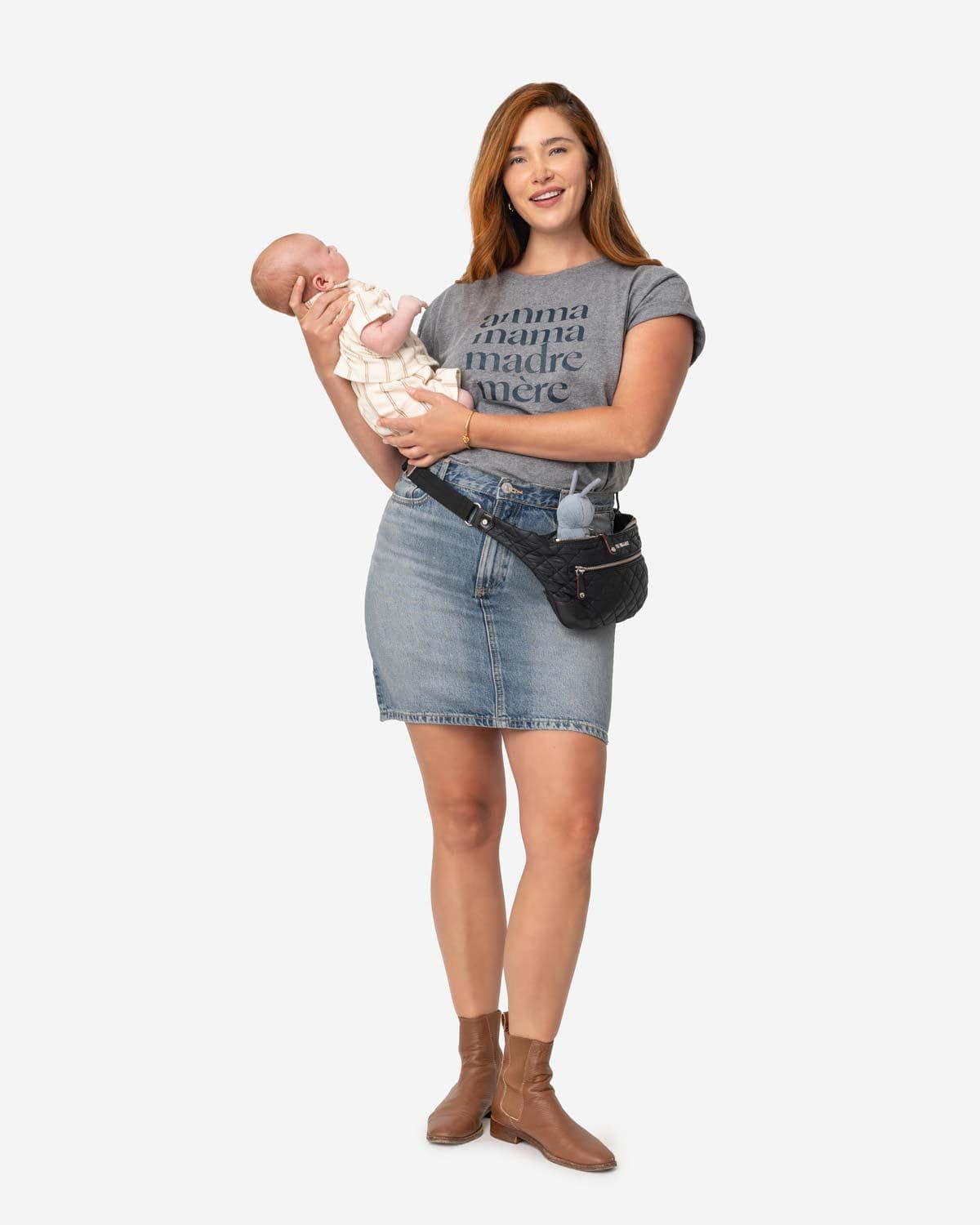 Young mom with long red hair holding her newborn baby while wearing a fanny pack, denim skirt and a heathered grey tee shirt that reads Amma, Mama, Madre, Mere in size XL