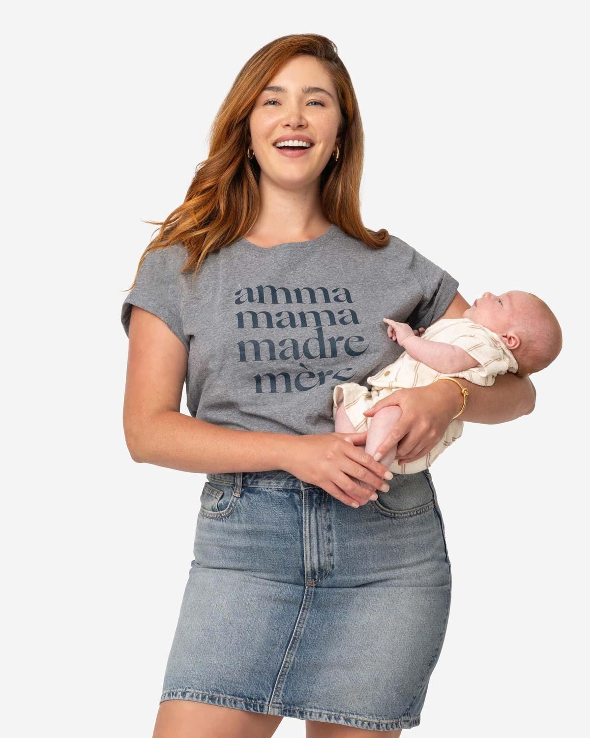 Young mom with long red hair holding her newborn baby while wearing denim skirt and a heathered grey tee shirt that reads Amma, Mama, Madre, Mere in size XL on a white background