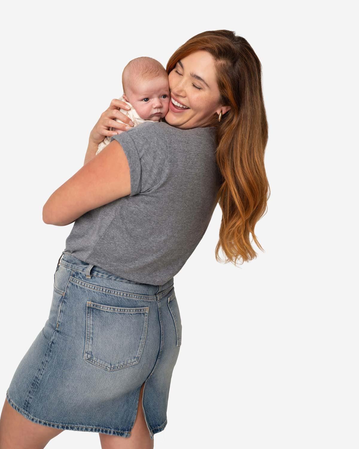 Back view of a young mom with long red hair holding her newborn baby while wearing denim skirt and a heathered grey tee shirt that reads Amma, Mama, Madre, Mere in size XL on a white background