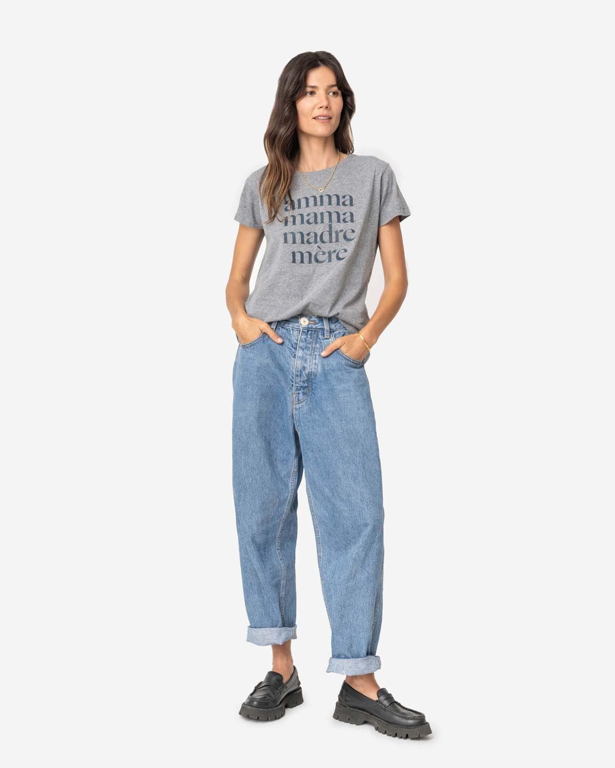 Young mom with long brown hair wearing loose jeans and a heathered grey tee shirt that reads Amma, Mama, Madre, Mere in size S on a white background