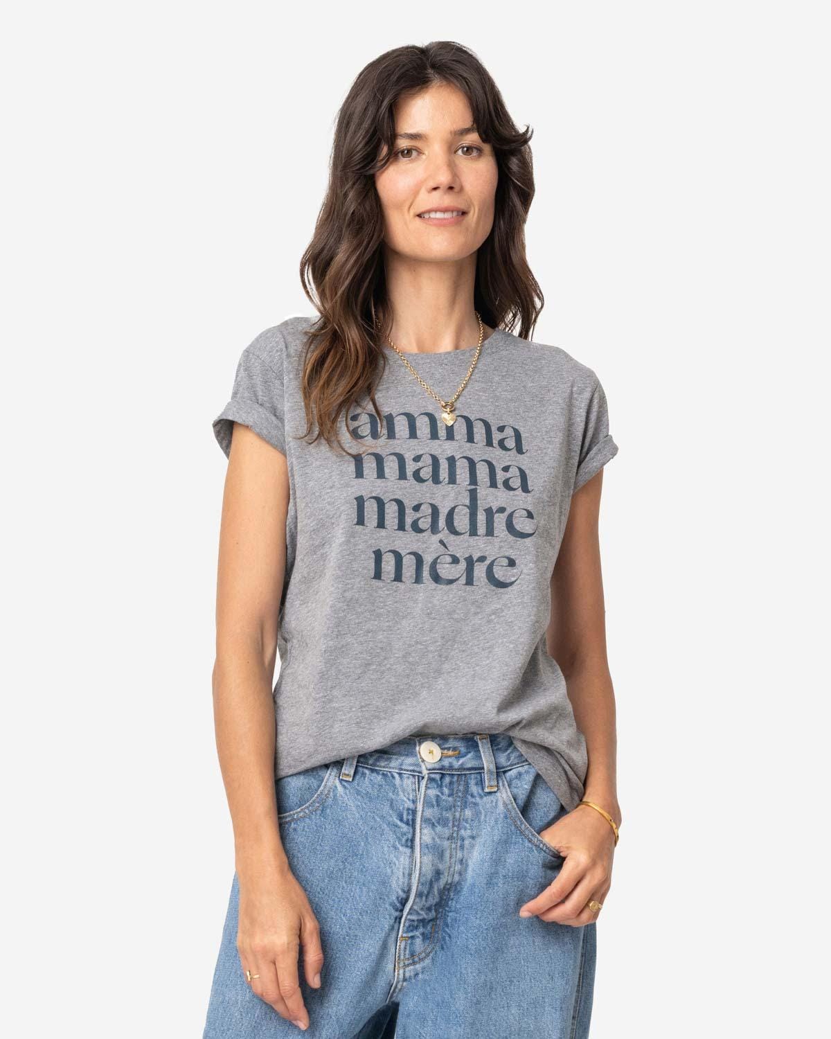 Young mom with long brown hair wearing loose jeans and a heathered grey tee shirt that reads Amma, Mama, Madre, Mere in size S on a white background