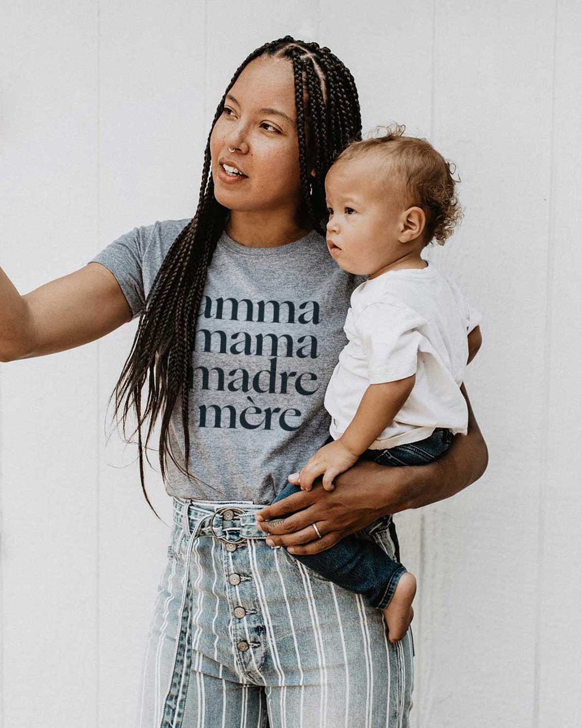 Young Black mom with long brown hair in braids pointing at something in the distance while holding her baby son and wearing a heathered grey tee shirt that reads Amma, Mama, Madre, Mere