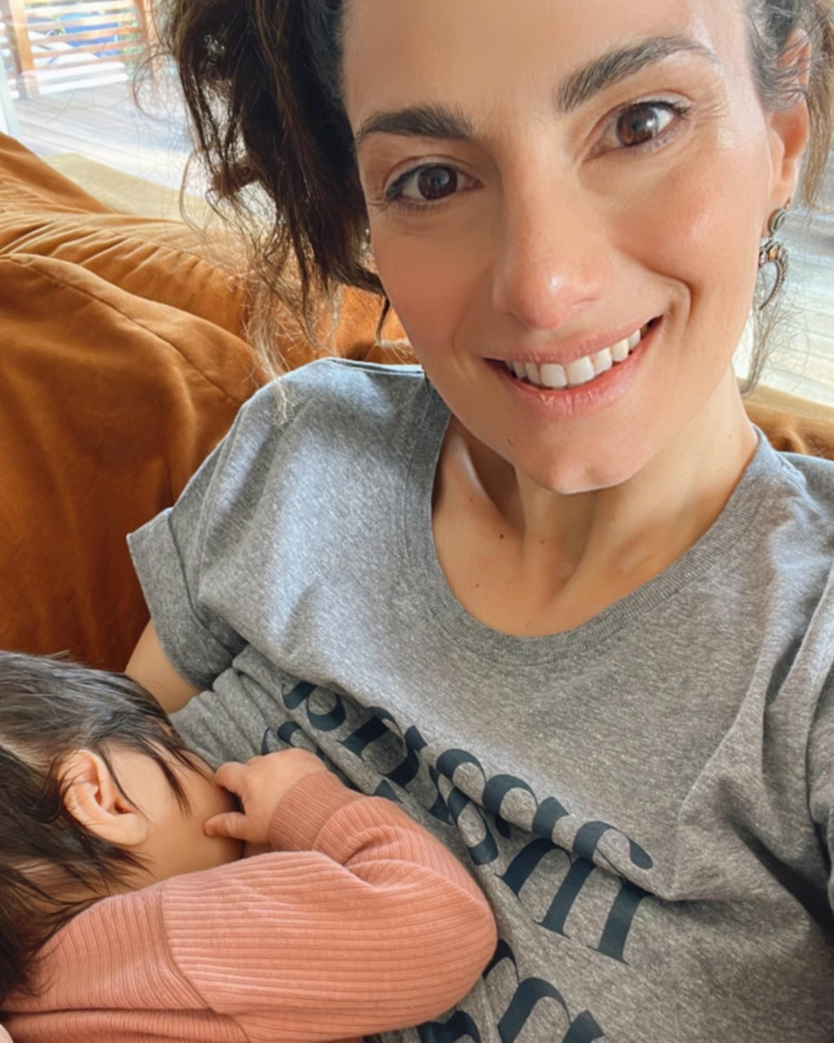 Mom with brown hair nursing her baby on a sofa and wearing a heathered grey tee shirt that reads Amma, Mama, Madre, Mere