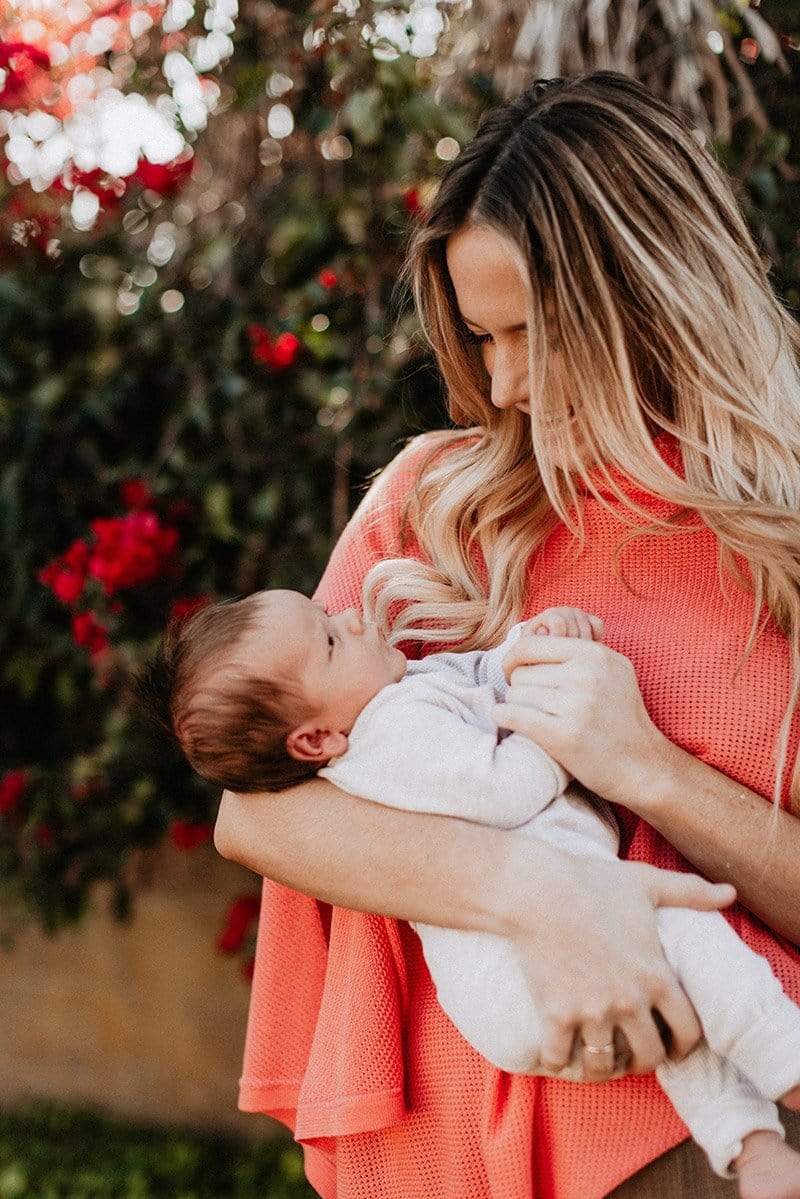 Blond woman in coral knit Cocoon nursing cover holding newborn baby