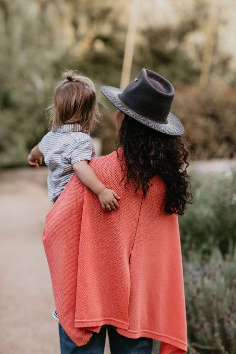 Back view of Satya Twena wearing coral knit Cocoon nursing cover styled as cape and holding toddler